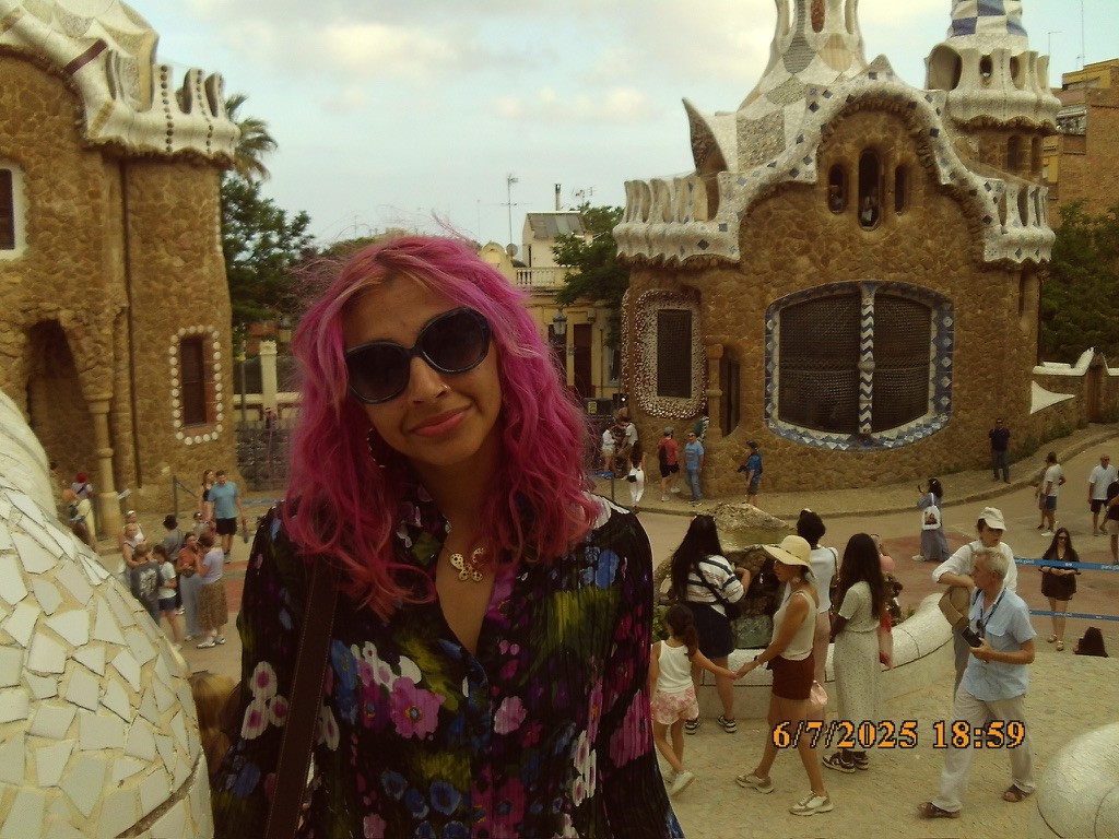 ID: A headshot of Avi Kumar. Description: A woman with pink hair and sunglasses, wearing a black button down and smiling at the camera.